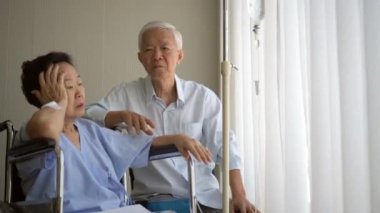 Asyalı yaşlı çift tekerlekli sandalye yaşı ve sağlık promblemi üzerine hastanede konuşan eşi destekliyor.