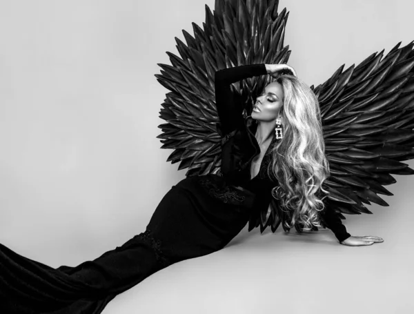 Stunning blonde woman in elegant long black dress and big wings is posing in studio. Luxury evening fashion. Glamour fashion model. Woman with wings. Black Angel. Black and white photo.