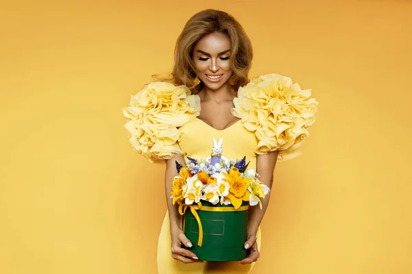 Beautiful happy woman in yellow spring dress is posing with Easter bouquet of flowers on a yellow background in studio. Easter concept.