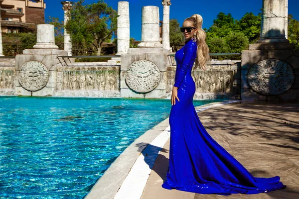 ブルーの長い夜のガウンのドレスを着た魅力的でスタイリッシュなエレガントな女性は 豪華なリゾート屋外でプールの近くにポーズをとっています — ストック写真