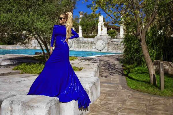 ブルーの長い夜のガウンのドレスを着た魅力的でスタイリッシュなエレガントな女性は 豪華なリゾート屋外でプールの近くにポーズをとっています — ストック写真