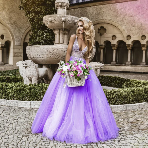 stock image Glamour, stylish elegant woman in lilac long evening gown dress is posing near the fountain in luxury resort outdoor.