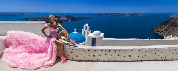 グラマー ピンクの長い結婚式のドレスのスタイリッシュなエレガントな花嫁の女性はサントリーニ島のFiraの3つの鐘の教会の近くにポーズをとっています — ストック写真