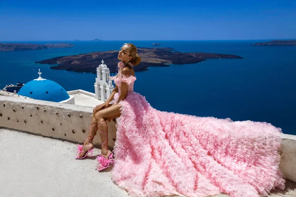 グラマー ピンクの長い結婚式のドレスのスタイリッシュなエレガントな花嫁の女性はサントリーニ島のFiraの3つの鐘の教会の近くにポーズをとっています — ストック写真
