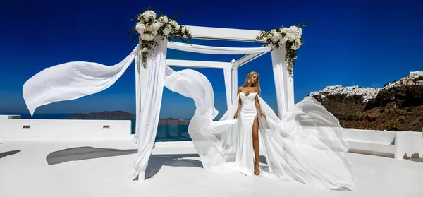 白の長いなびく結婚式のドレスでスタイリッシュなエレガントな花嫁の女性はサントリーニ島のFiraの結婚式のエリアでポーズをとっています — ストック写真