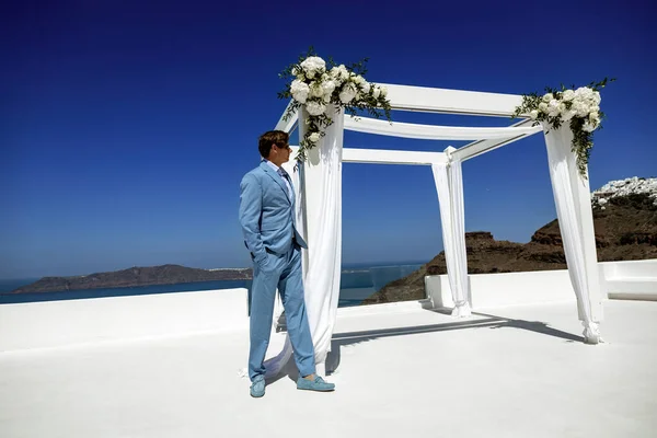 Elegant handsome groom in blue suit is posing on wedding area in Santorini. Groom. Engagement and wedding in Santorini island of Greece. Greek wedding