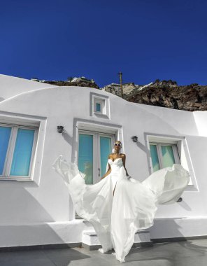 Beyaz, uzun, kanat çırpan gelinlik içinde zarif bir kadın Santorini manzarasında beyaz binalar ve güzel pembe çiçeklerle poz veriyor.