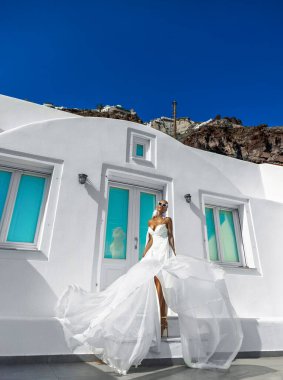 Beyaz, uzun, kanat çırpan gelinlik içinde zarif bir kadın Santorini manzarasında beyaz binalar ve güzel pembe çiçeklerle poz veriyor.