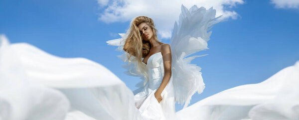 Beautiful sexy blonde woman in white long elegant dress and angel wings is sensually posing against the heaven on summer day. Angel woman.