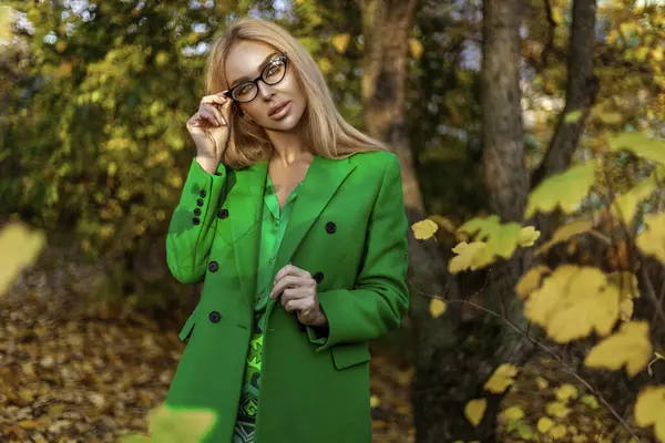 Beautiful woman wearing a green coat and elegant clothing is posing outdoors on a sunny autumn day.