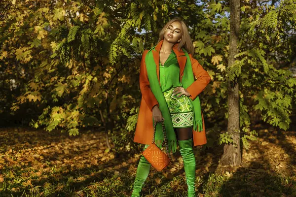 Turuncu ceketli ve zarif yeşil elbiseli güzel bir kadın güneşli bir sonbahar gününde dışarıda poz veriyor..