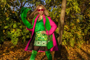 Pembe zarif bir ceket ve yeşil eşarp giyen güzel bir kadın güneşli bir sonbahar gününde dışarıda poz veriyor..