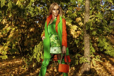 Turuncu ceketli ve zarif yeşil elbiseli güzel bir kadın güneşli bir sonbahar gününde dışarıda poz veriyor..