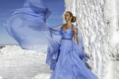 Mavi kanat çırpan uzun elbiseler giyen zarif bir kadın ve güneşli bir günde dağlarda poz veriyor. Kar Kraliçesi.