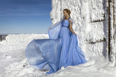 Mavi kanat çırpan uzun elbiseler giyen zarif bir kadın ve güneşli bir günde dağlarda poz veriyor. Kar Kraliçesi.