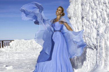 Mavi kanat çırpan uzun elbiseler giyen zarif bir kadın ve güneşli bir günde dağlarda poz veriyor. Kar Kraliçesi.