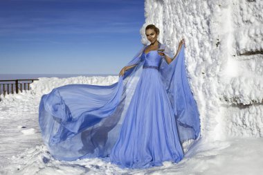 Mavi kanat çırpan uzun elbiseler giyen zarif bir kadın ve güneşli bir günde dağlarda poz veriyor. Kar Kraliçesi.