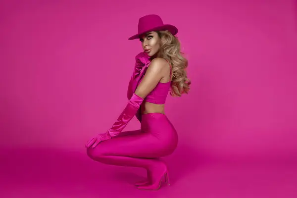 The beautiful model is wearing sexy pink shiny leggings, ankle boots, an elegant fuchsia top and a pink hat and is posing against a pink background. Spring model on a pink background.