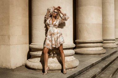 Çiçekli bir elbise ve seksi topuklu ayakkabılar giyen zarif, şık bir kadın yaz günü şehirde şehvetle poz verir. Açık oturum.