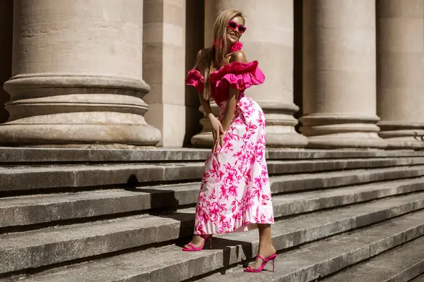 ピンクの花のドレスとセクシーなハイヒールのエレガントでスタイリッシュな女性は 晴れた夏の日に街を感動的に歩いています アウトドアシュート ロイヤリティフリーのストック写真