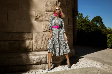 Şık, şık bir kadın, kadınsı çiçekli bir elbise ve seksi topuklu ayakkabılar içinde güneşli bir yaz gününde şehrimizde şehvetli bir şekilde yürüyor. Açık hava çekimi.