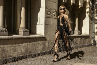 Siyah Macrame elbiseli ve yüksek topuklu ayakkabılı seksi sarışın manken güneşli bir yaz gününde şehrimizde şehvetle yürüyor. Açık hava çekimi..
