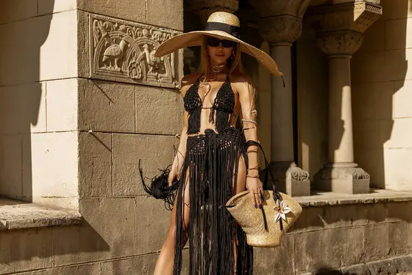Siyah Macrame elbisesi içinde şık, şık bir kadın, seksi topuklu ayakkabılar ve büyük bir şapka güneşli bir yaz gününde şehrimizde şehvetle yürüyor. Açık hava çekimi..