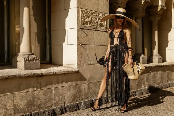 Siyah Macrame elbisesi içinde şık, şık bir kadın, seksi topuklu ayakkabılar ve büyük bir şapka güneşli bir yaz gününde şehrimizde şehvetle yürüyor. Açık hava çekimi..