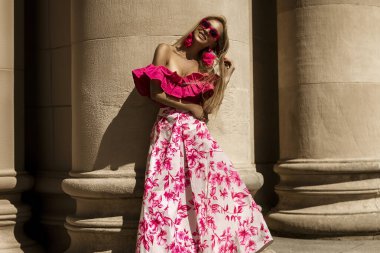 Pembe çiçekli elbiseler ve seksi topuklu ayakkabılar giyen zarif, şık bir kadın güneşli bir yaz gününde şehrimizde şehvetli bir şekilde yürüyor. Açık hava çekimi..