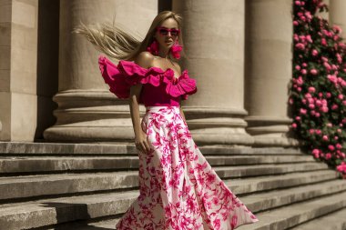 Pembe çiçekli elbiseler ve seksi topuklu ayakkabılar giyen zarif, şık bir kadın güneşli bir yaz gününde şehrimizde şehvetli bir şekilde yürüyor. Açık hava çekimi..