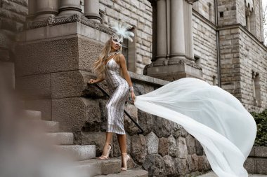 Şık lüks akşam modası. Şato bahçesinde gümüş pullu gece elbisesi ve tüy karnaval maskesi takan alımlı zarif bir kadın poz veriyor. Parti elbisesi içindeki kadın model. Vogue. Özel tasarım.