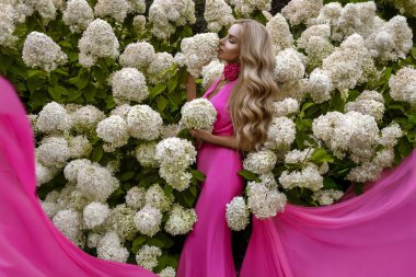 Dalgalı pembe elbiseli zarif sarışın kadın. Çiçekli, rüya gibi bahar çiçekleriyle süslü ortancaları var. Zarafet, kadınlık, romantizm ve botanik ilham.