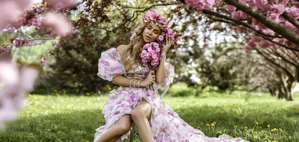 Çiçekli, pembe bir elbise ve çiçek desenli bir taç giymiş zarif sarışın bir manken çiçek açan bir bahçede oturmuş romantik bahar havası yayıyor, kadınlık ve doğal güzelliği etrafında çiçeklerle dolanıyor..