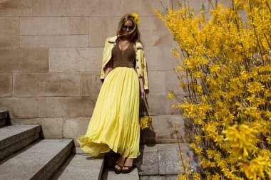 Akan sarı Maxi elbiseli zarif kadın güneşli bir yaz gününde şehirde sarı fındık çiçeklerinin yanında poz verir..