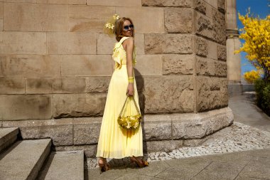 Akan sarı Maxi elbiseli zarif kadın güneşli bir yaz gününde şehirde sarı fındık çiçeklerinin yanında poz verir..