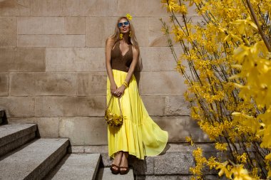 Akan sarı Maxi elbiseli zarif kadın güneşli bir yaz gününde şehirde sarı fındık çiçeklerinin yanında poz verir..