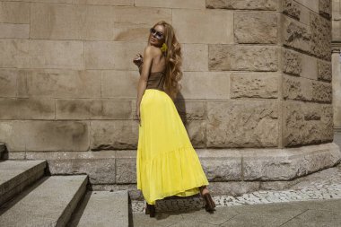 Akan sarı Maxi elbiseli zarif kadın güneşli bir yaz gününde şehirde sarı fındık çiçeklerinin yanında poz verir..