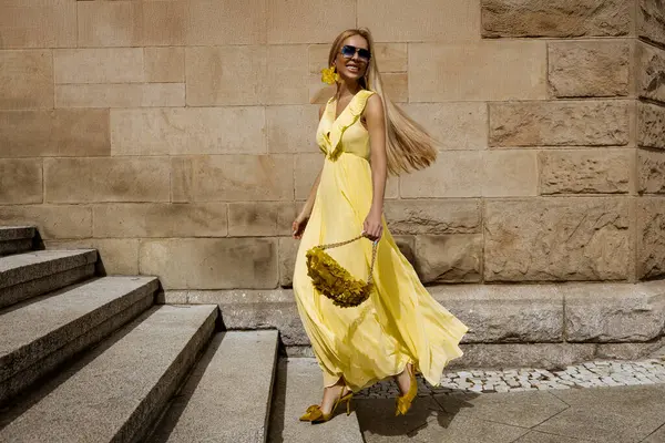 Akan sarı Maxi elbiseli zarif kadın güneşli bir yaz gününde şehirde sarı fındık çiçeklerinin yanında poz verir..