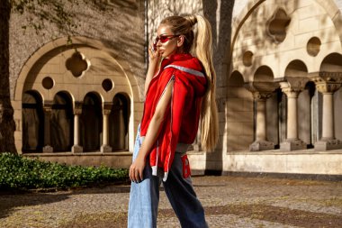 Güneşli bir yaz gününde şehirde kırmızı ceketli, güneş gözlüklü ve kot pantolonlu genç ve şık bir kadınla yaz modası. Denim modası.