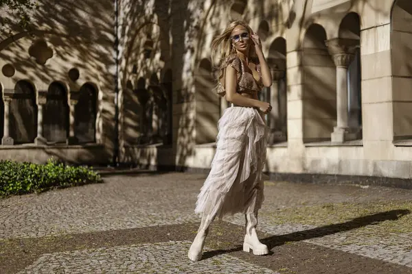 Tulle bej uzun etekli zarif bir kadın ve yaz güneşinde şehirde poz veren şık bir pullu bluz. Yaz şehir modası.