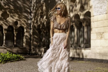 Tulle bej uzun etekli zarif bir kadın ve yaz güneşinde şehirde poz veren şık bir pullu bluz. Yaz şehir modası.