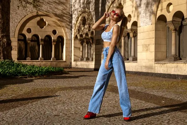 Yazlık kot pantolonlu, seksi bluzlu ve güneşli yaz gününde şehirde poz veren güneş gözlüklü bir kadın. Kot pantolon moda..