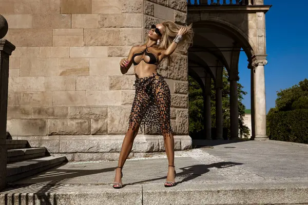 Havalı bir bikini ve lüks plaj eteğiyle mükemmel bronzlaşmış vücudu olan seksi sarışın manken güneşli bir yaz gününde şehirde şehvetli pozlar veriyor. Açık oturum.