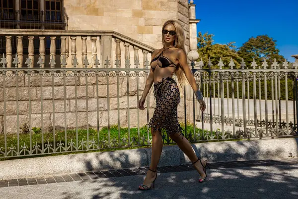 Şık bikinili, mükemmel bronzlaşmış vücudu olan ve kristalli lüks eteği olan seksi sarışın manken güneşli bir yaz gününde şehrimizde şehvetle yürüyor. Açık oturum.