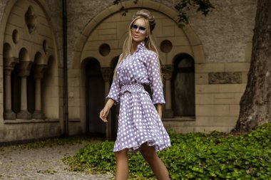 Uzun puantiyeli yaz elbisesi ve şık topuklu ayakkabılı zarif bir kadın güneşli bir yaz gününde şehirde şehvetli pozlar veriyor. Yaz Moda..