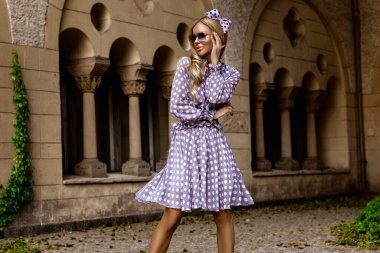 Uzun puantiyeli yaz elbisesi ve şık topuklu ayakkabılı zarif bir kadın güneşli bir yaz gününde şehirde şehvetli pozlar veriyor. Yaz Moda..