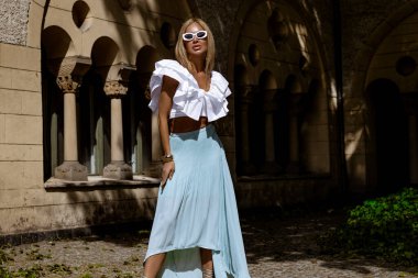 Uzun sarı saçlı, mavi etekli ve dalgalı bluzlu zarif manken güneşli bir yaz gününde şehrimizde şehvetli pozlar veriyor. Yaz modası.