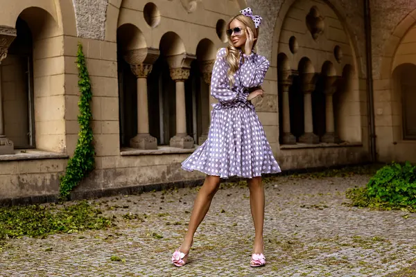 Uzun puantiyeli yaz elbisesi ve şık topuklu ayakkabılı zarif bir kadın güneşli bir yaz gününde şehirde şehvetli pozlar veriyor. Yaz Moda..