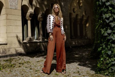 Şık bir yaz tulumu ve boho ceketiyle şık bir kadın model güneşli bir yaz gününde şehirde şehvetli pozlar veriyor. Yaz modası.