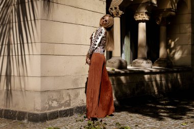 Şık bir yaz tulumu ve boho ceketiyle şık bir kadın model güneşli bir yaz gününde şehirde şehvetli pozlar veriyor. Yaz modası.